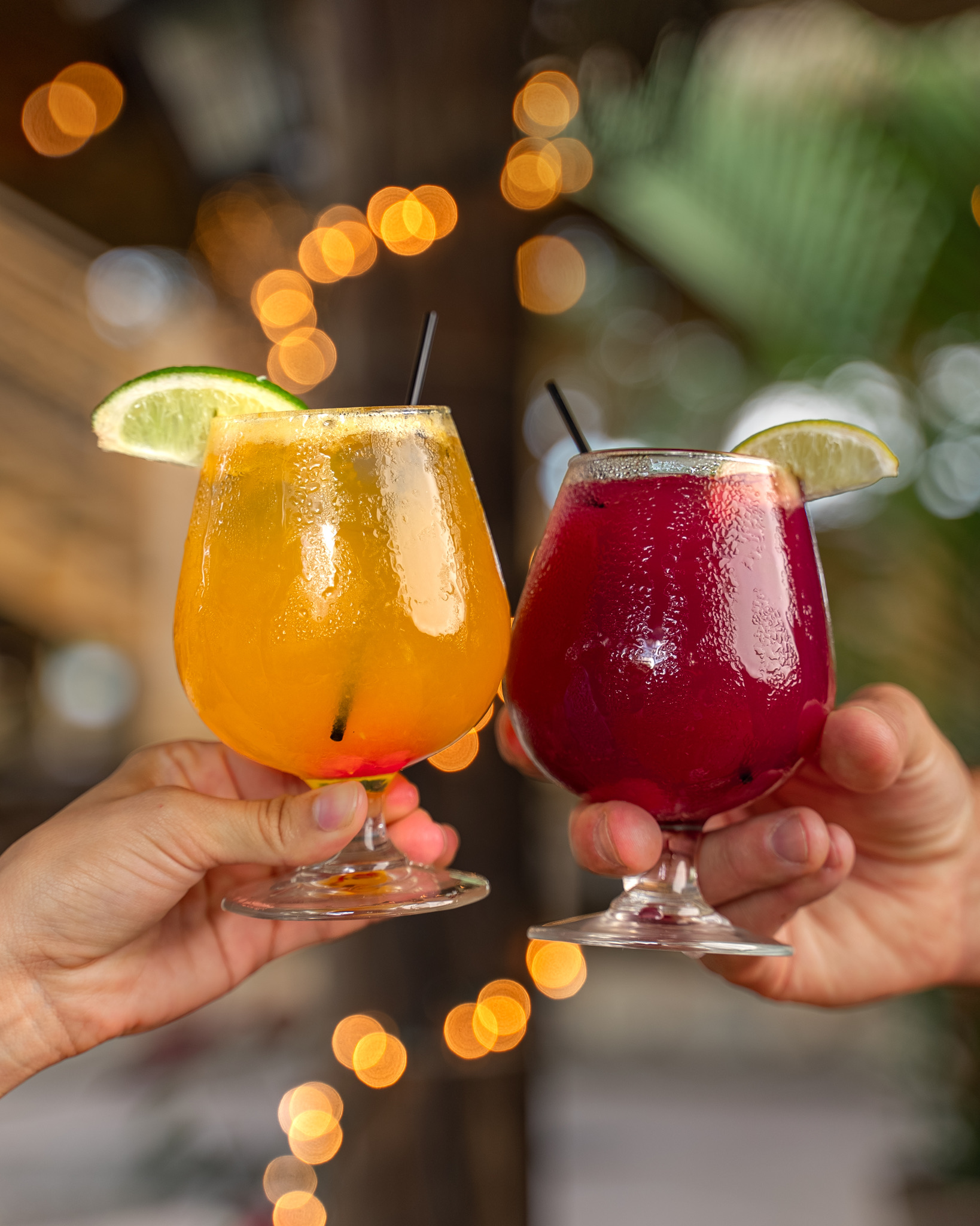 Two hands toasting with orange and red cocktails, garnished with lime, against a blurred light backdrop.