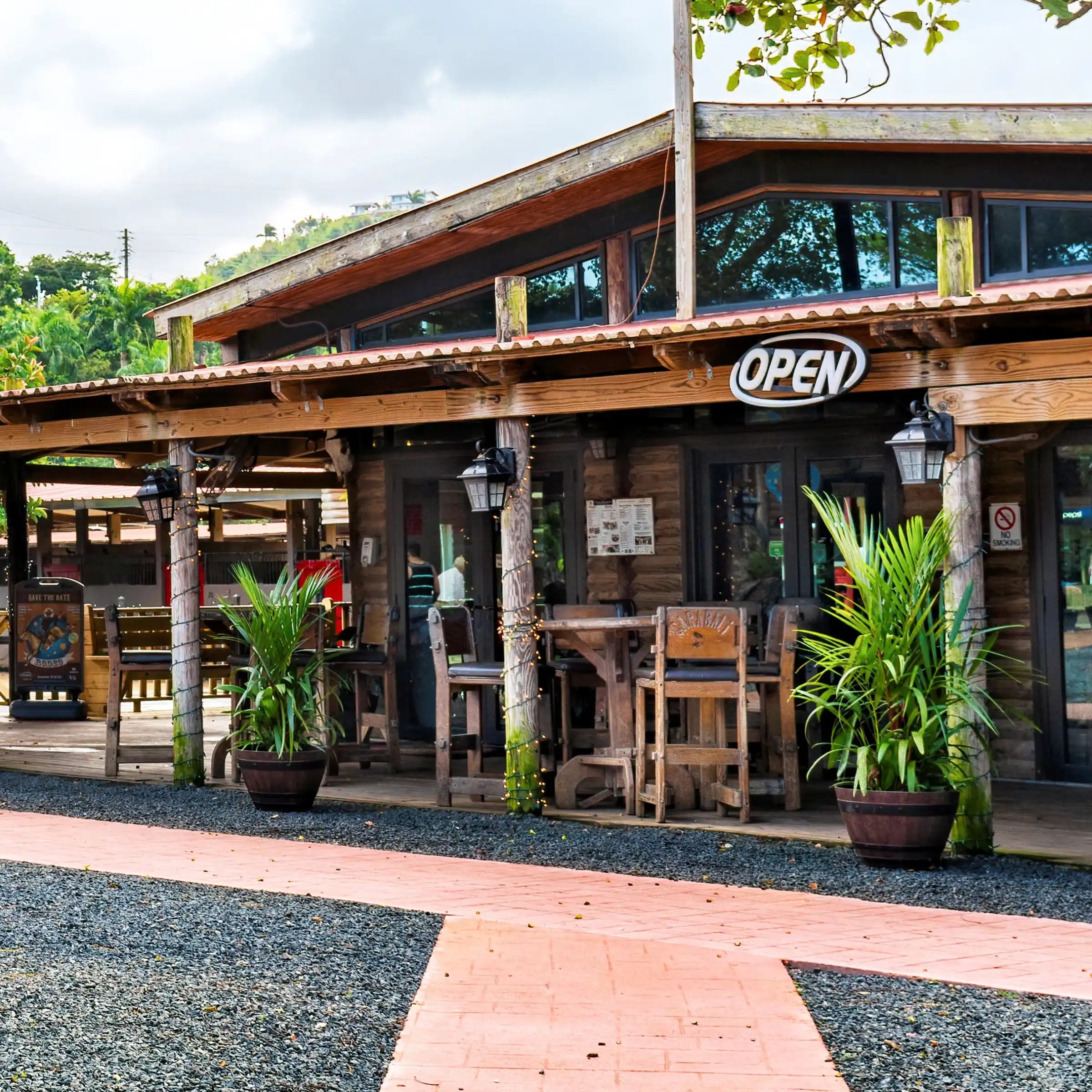 Outdoor view of a rustic café with an 'Open' sign, wooden tables, and potted plants.