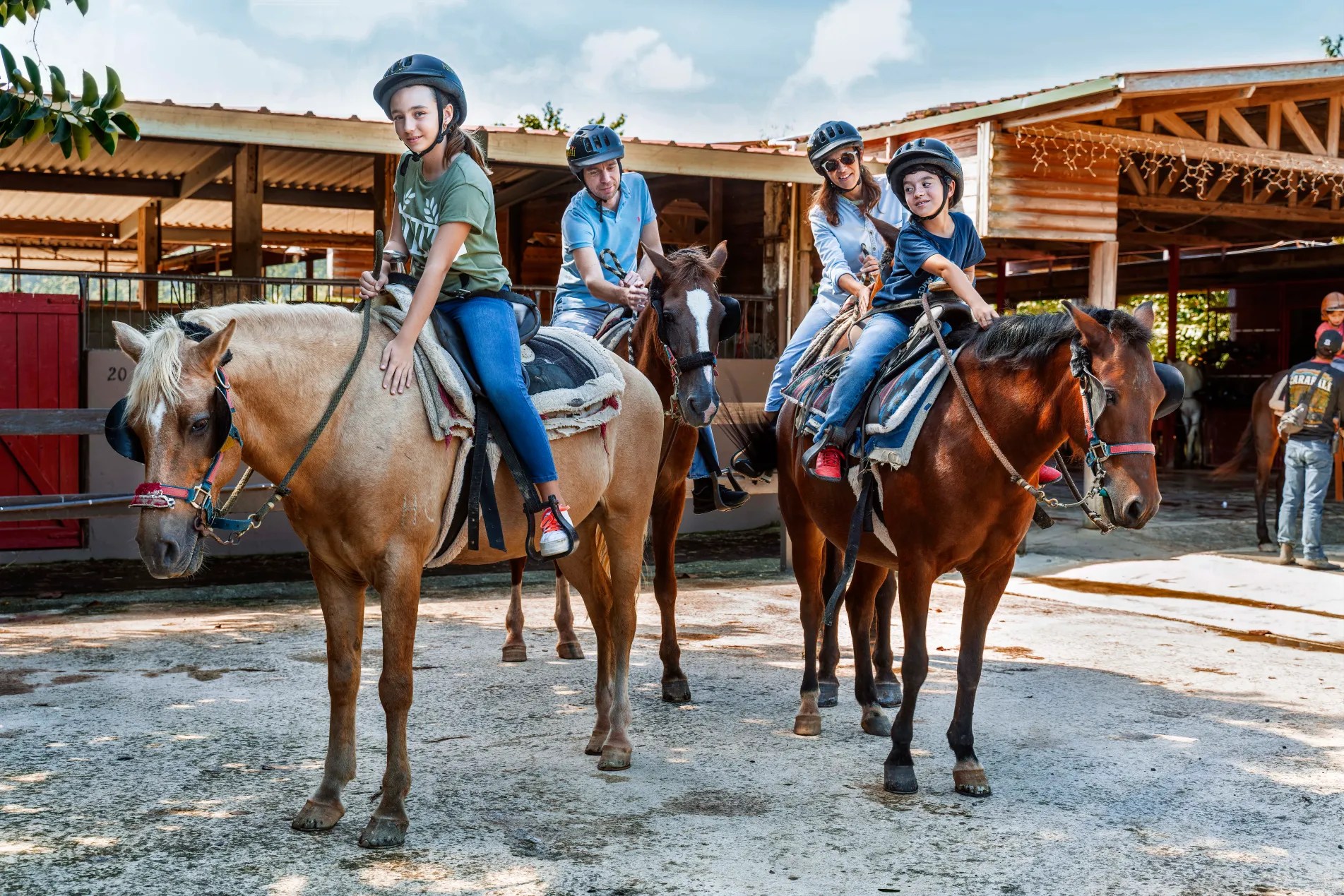 Four people wearing helmets on horses outside a stable.