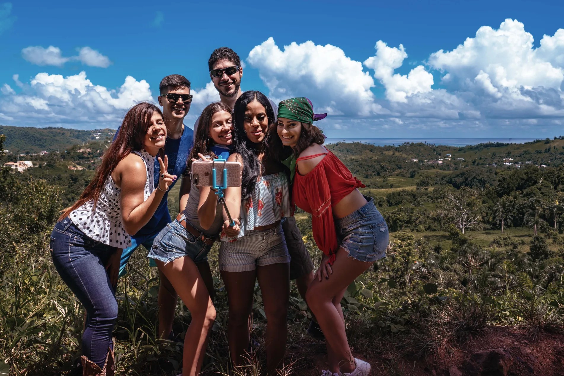 Group of six friends taking a selfie with a scenic landscape in the background on a sunny day.