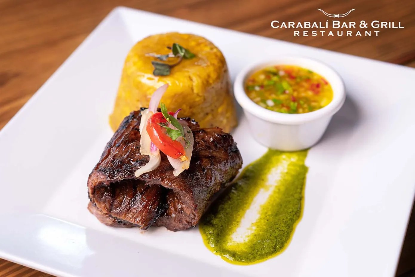 Steak with chimichurri, mofongo, and a dipping sauce on a white plate.