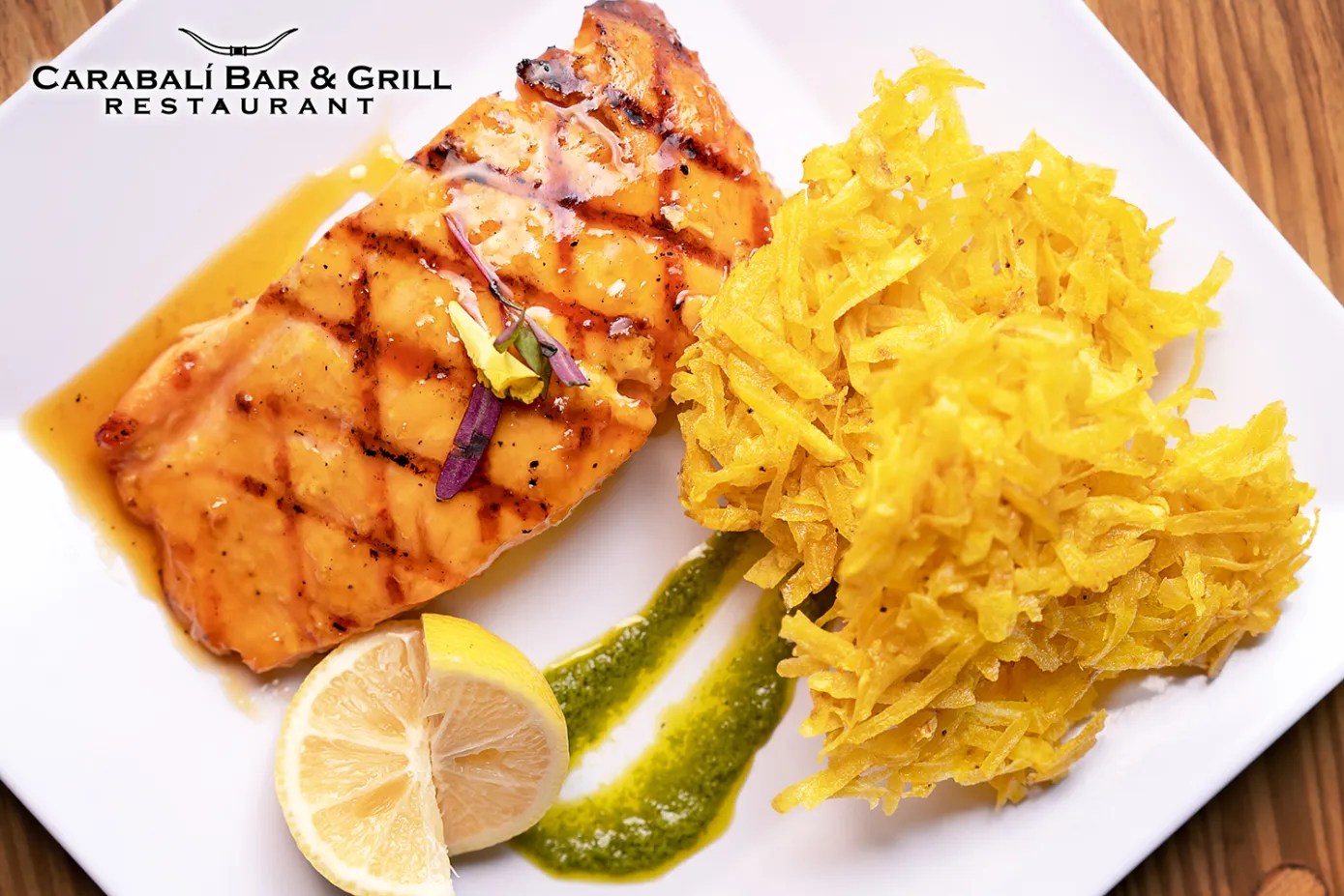 Grilled salmon with shredded yellow vegetables, lemon slice, and green sauce on a white plate.