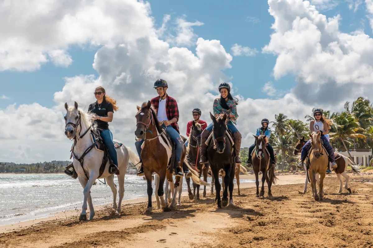 Group of people riding horses on a sandy beach under a partly cloudy sky.