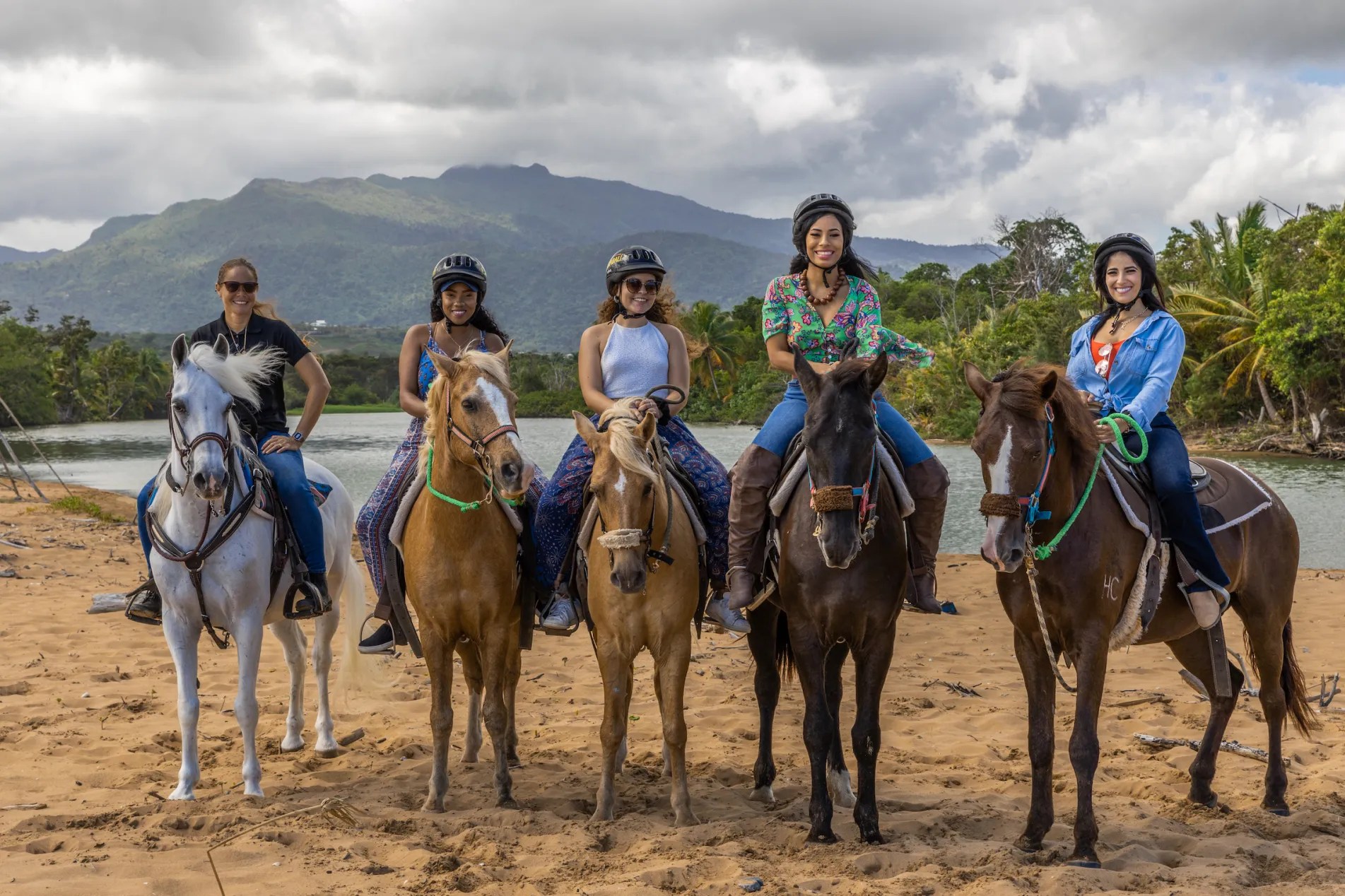 Five people on horseback on a sandy beach with mountains in the background.