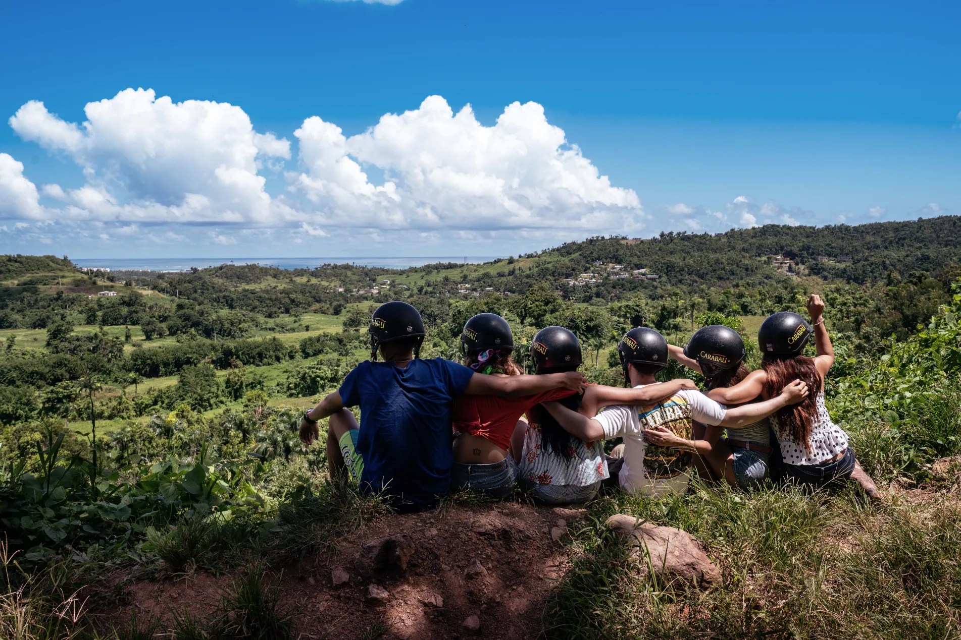 Group of people with helmets sitting on a hill, overlooking a scenic landscape with clouds.